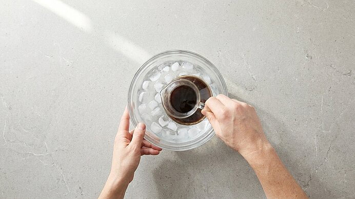 Zwei Hände halten eine Glasschale mit Eis, während aus einer kleinen Tasse konzentrierter Espresso über das Eis gegossen wird.