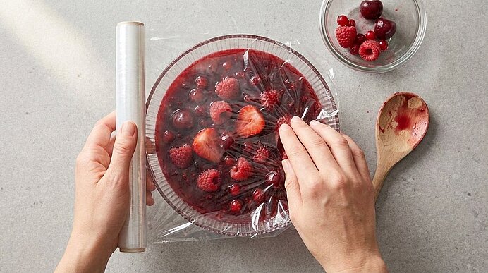 Zwei Hände spannen Frischhaltefolie über eine Glasschale mit roten Beeren und Gelee; daneben eine kleine Schüssel mit Himbeeren und Kirschen sowie ein Holzlöffel.