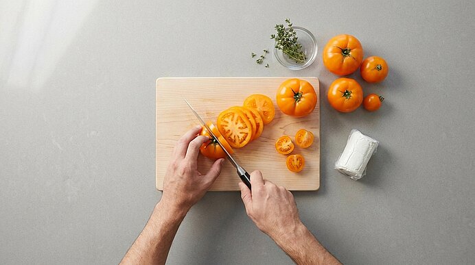 Hände schneiden mit einem Messer orangefarbene Tomaten auf einem Holz-Schneidebrett; mehrere ganze und geschnittene Tomaten liegen daneben.
