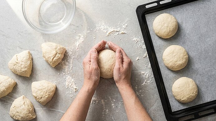 Zwei Hände formen ein Teigstück auf der Arbeitsplatte. Daneben liegt ein Backblech mit Brötchen.