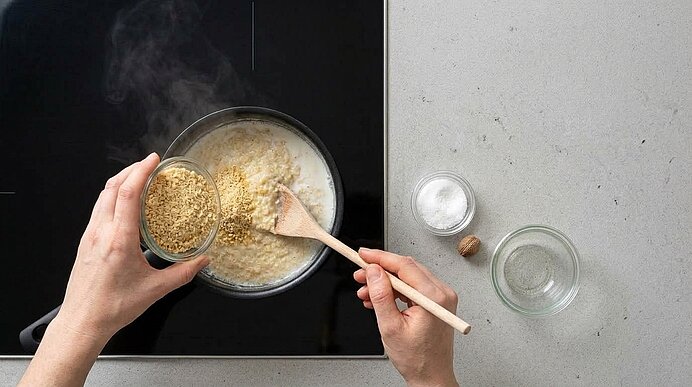 Hände rühren Riebelmais aus einer Schale mit einem Holzlöffel in die heiße Milch im Topf auf dem Kochfeld.