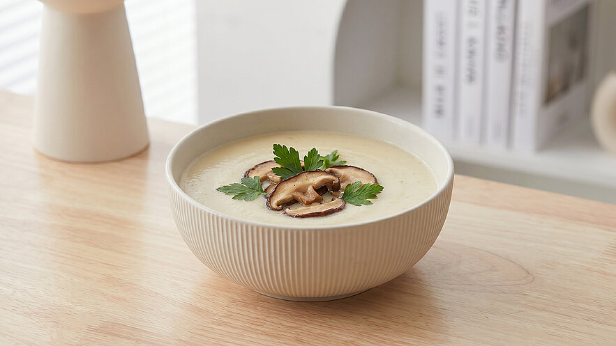 Eine schlichte, weiße Schüssel mit cremiger Suppe steht auf einem Holztisch. Die Suppe ist mit einigen Scheiben Pilzen und frischer Petersilie garniert. Im Hintergrund sind verschwommen Bücher und eine Vase sichtbar.