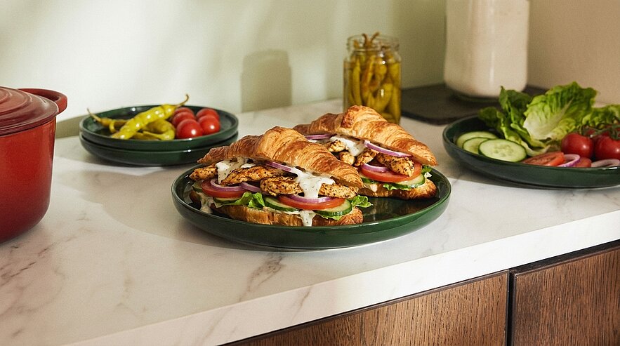 Auf dem Bild sind mehrere üppige Croissant-Sandwiches auf einem grünen Teller zu sehen, garniert mit frischem Gemüse wie Tomaten, Gurken und Salat. Im Hintergrund sind weitere Schalen mit frischen Lebensmitteln und ein roter Topf sichtbar.