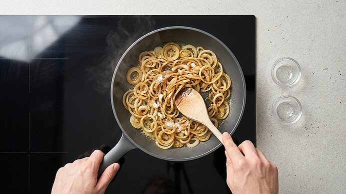 Zwei Hände rühren mit einem Holzlöffel angebräunte Zwiebelringe in einer Pfanne auf dem Herd, Dampf steigt auf.