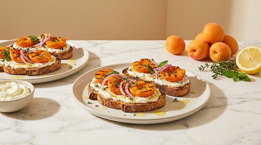 Drei Scheiben Brot mit Ricottafrischkäse, gegrillten Aprikotenhälften, roten Zwiebelringen und Kräutern, beträufelt mit Olivenöl; im Hintergrund frische Aprikosen und eine Zitronenhälfte.