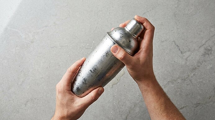 Zwei Hände schütteln einen nassen Metallcocktailshaker; Tropfen spritzen von ihm auf eine helle Arbeitsfläche.