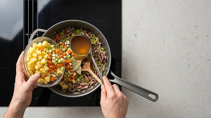 Hände füllen gewürfelte Kartoffeln und Brühe in einen Topf mit Karotten, Lauch und Speck; ein Holzlöffel rührt auf dem Herd.