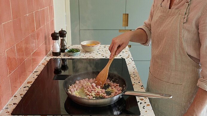 Oberkörper einer Person in Schürze rührt mit Holzlöffel in einer Pfanne auf dem Herd; darin braten gewürfelter Speck und Zwiebelstücke.
