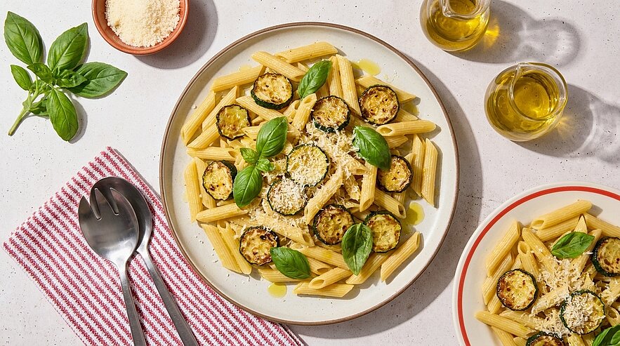 Teller mit Penne-Nudeln, gegrillten Zucchinischeiben, frischen Basilikumblättern, geriebenem Parmesan und Olivenöl.