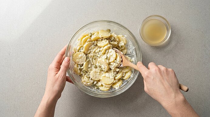 Zwei Hände rühren mit einem Holzlöffel in einer Glasschüssel Kartoffelsalat aus dünn geschnittenen Kartoffeln, Zwiebeln und cremigem Dressing; daneben steht eine kleine Schale mit zusätzlichem Dressing.