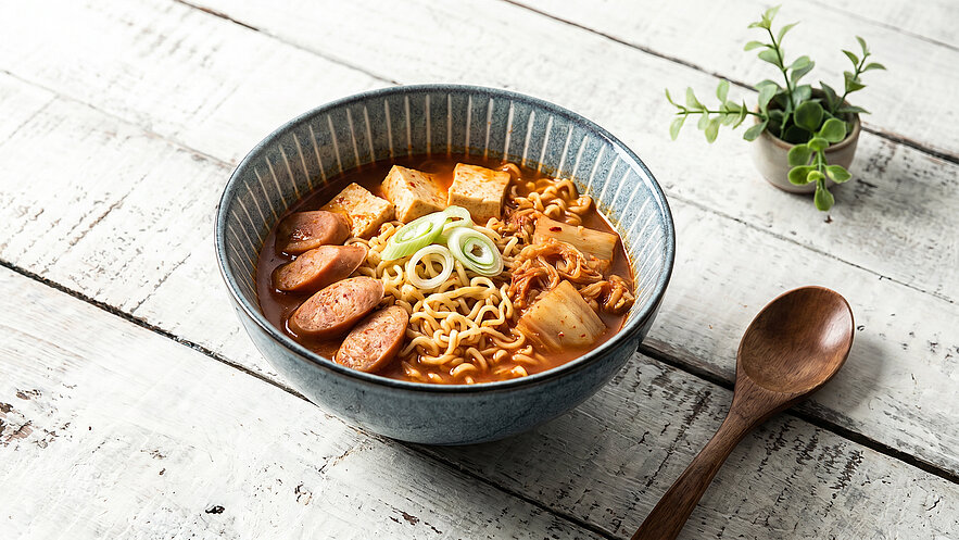 Eine Schüssel mit Nudelsuppe, die gekochte Nudeln, Würstchen, Tofu und Frühlingszwiebeln enthält. Im Hintergrund steht eine kleine Pflanze. Die Schüssel hat ein elegantes, blaues Design und die Suppe hat eine appetitliche, würzige Farbe.