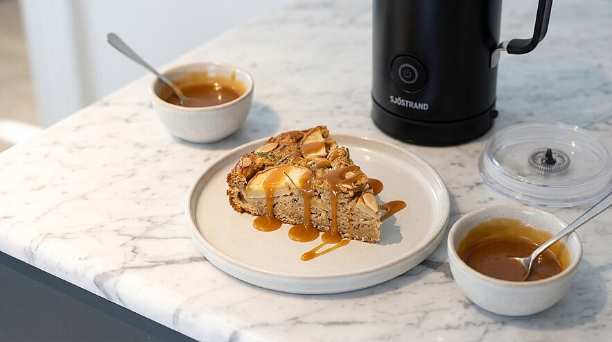 Stück Kuchen mit Karamellsauce auf einem weißen Teller auf Marmorplatte, daneben zwei kleine Schälchen mit Sauce und ein schwarzes elektrisches Gerät im Hintergrund.
