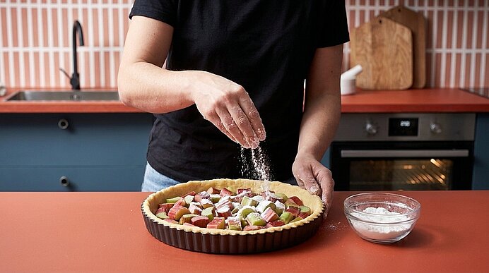 Hände einer Person bestreuen einen rohen Obstkuchen in einer Tarteform mit Puderzucker; neben der Form steht eine Schüssel mit Zucker auf der Küchenarbeitsfläche.