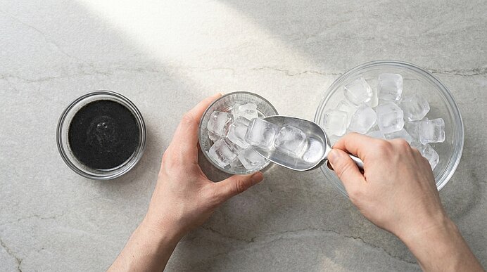 Von oben: Hände schaufeln mit einer Eiskelle Eiswürfel aus einer Schüssel in ein Glas.