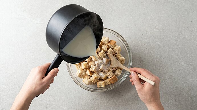 Hände gießen warme Milch aus einem kleinen schwarzen Topf über gewürfeltes Brot in einer Glasschüssel; eine Hand hält einen Holzspatel zum Verrühren.