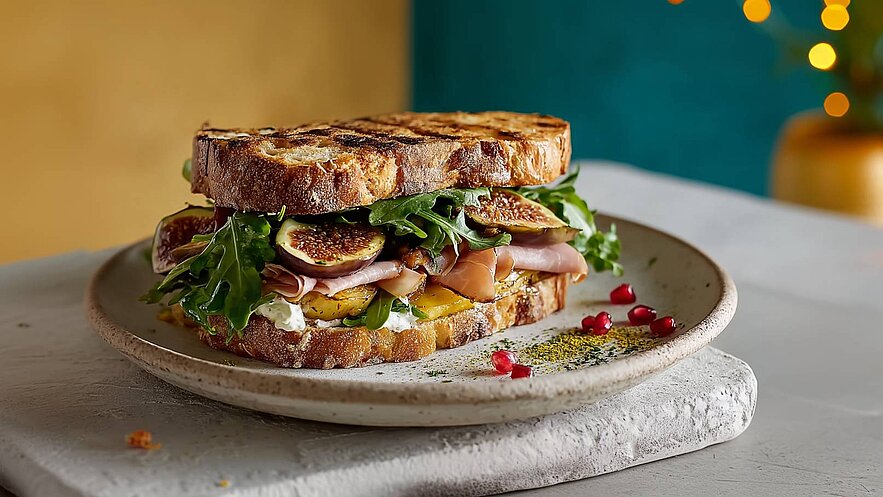 Ein rustikales Sandwich besteht aus geröstetem Brot, gefüllt mit frischen Feigen, Rucola, Schinken und einer cremigen Schicht. Es liegt auf einem hellen Teller, umgeben von einigen Granatapfelkernen und Gewürzen. Der Hintergrund ist in sanften Farben gehalten.