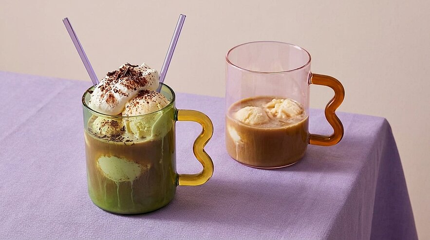 Zwei transparente Gläser mit dekorativen Griffen: links grüner Schichtdrink mit Sahne, Kakaostreuseln und zwei Strohhalmen; rechts brauner Kaffee mit einer Kugel Vanilleeis.