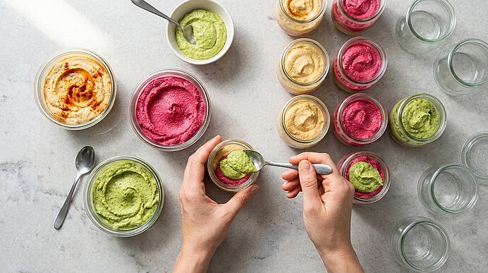 Zwei Hände füllen dreifarbigen Hummus schichtweise mit einem Löffel in kleine Gläser auf der hellgrauen Arbeitsplatte.