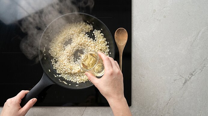 Hände rühren Reis in einer Pfanne auf dem Herd, während aus einem kleinen Glas Flüssigkeit hinzugegossen wird; Dampf steigt auf.