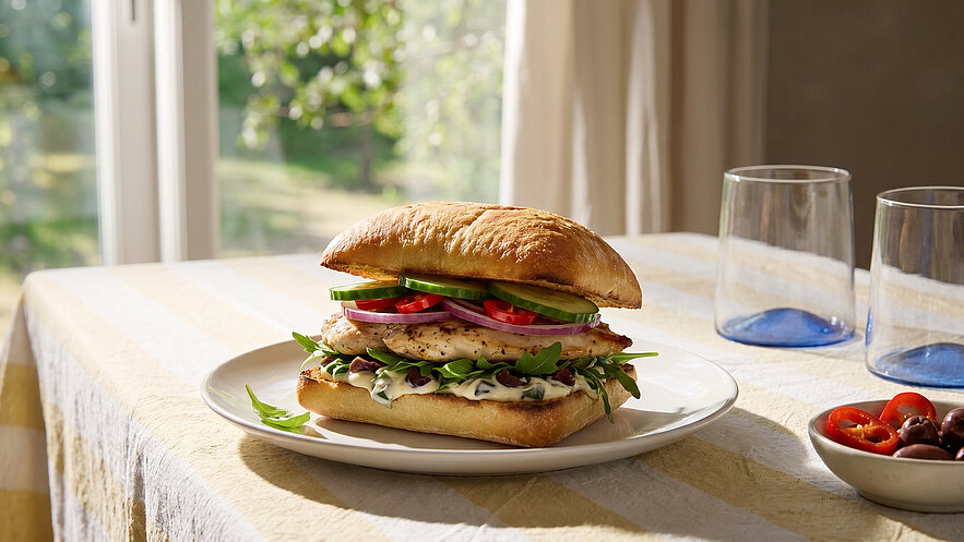 Ein frisch zubereiteter Sandwich auf einem weißen Teller, gefüllt mit Hähnchenbrust, frischem Gemüse wie Tomaten, Gurken und roten Zwiebeln, auf einem luftigen Brötchen. Im Hintergrund sind zwei Gläser und eine Schale mit Kirschtomaten sichtbar.