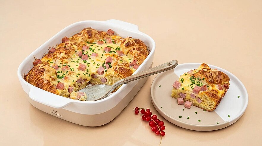 Auflaufform mit goldbraunem Croissant‑Auflauf, sichtbar Schinkenwürfel und geschmolzener Käse, mit Schnittlauch bestreut; eine Stückportion auf einem Teller daneben.