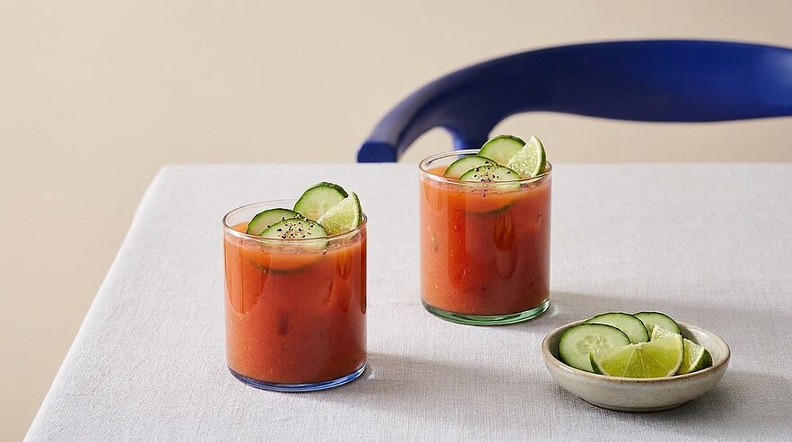 Zwei Gläser roter Tomatencocktail mit Gurkenscheiben und Limettenspalten als Garnitur; daneben eine kleine Schale mit zusätzlichen Gurken- und Limettenscheiben auf hellem Tischtuch.