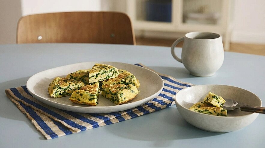 Teller mit in Stücke geschnittener Spinat-Frittata auf gestreiftem Tuch; daneben eine kleine Schale mit einem Stück und einer Gabel sowie eine Tasse auf dem Tisch.