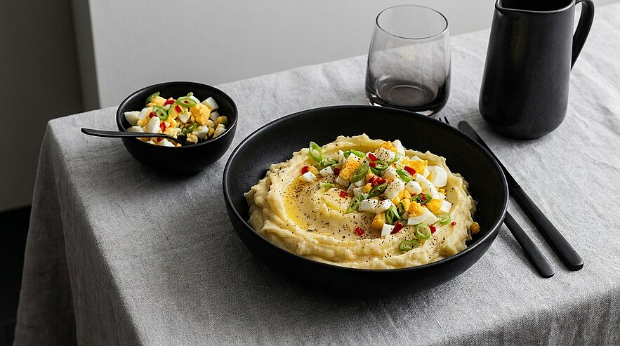 Schwarze Schale mit cremigem Kartoffelpüree, garniert mit gehacktem hartgekochtem Ei, Frühlingszwiebeln und Paprikawürfeln; daneben kleine Schale mit zusätzlicher Ei-Garnitur auf hellem Tuch.