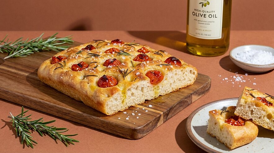Rechteckige Focaccia mit halben Kirschtomaten, Rosmarinzweigen und grobem Meersalz auf einem Holzbrett, zwei Stücke auf kleinem Teller.