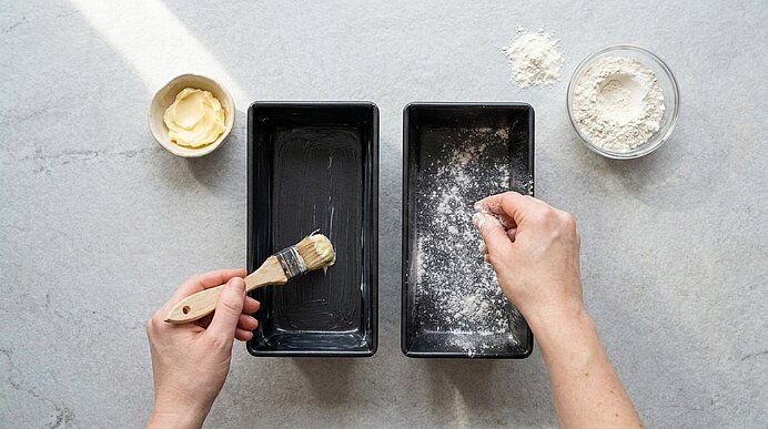 Zwei Kastenformen auf der Arbeitsfläche: links mit Butter eingepinselt, rechts mit Mehl bestäubt; Schälchen mit Butter und Mehl daneben.