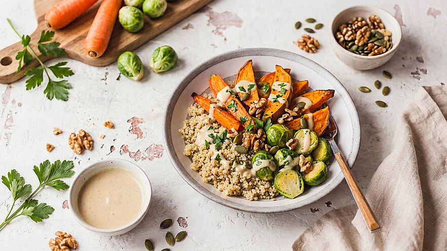 Eine Schüssel mit Quinoa als Basis, garniert mit gerösteten Süßkartoffeln, rohem Rosenkohl und Walnüssen. Darüber befindet sich eine cremige Sauce. Im Hintergrund sind frische Karotten und einige Kürbiskerne zu sehen.