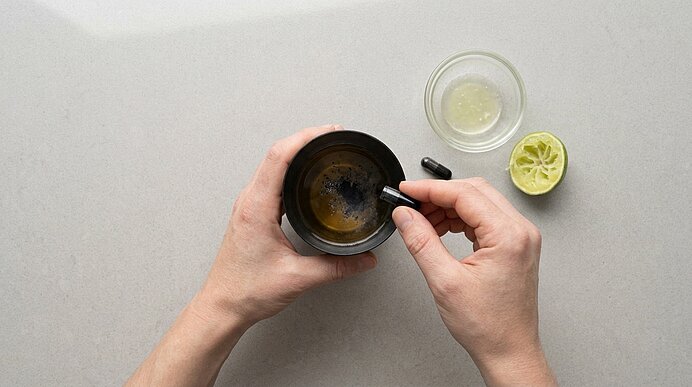 Zwei Hände geben Aktivkohlepulver aus einer Kapsel in einen Cocktailshaker. Daneben stehen Limettensaft und Limette.