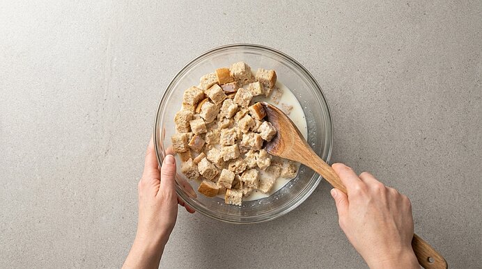 Zwei Hände rühren in einer Glasschüssel gewürfeltes Brot mit Milch mit einem Holzlöffel.