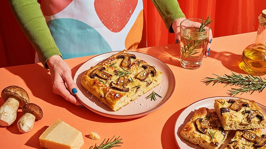 Hände halten einen goldbraunen, rechteckigen Focaccia-Fladen, belegt mit gebratenen Champignons und Rosmarin, in Stücke geschnitten.