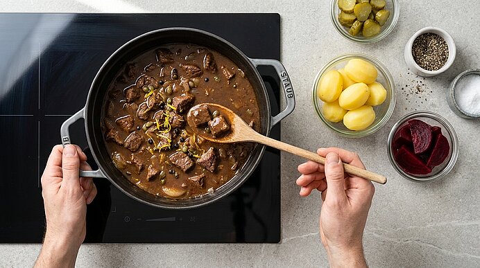 Hände rühren in einem schwarzen Schmortopf auf Cerankochfeld ein dunkles Rindergulasch mit Pilzen; daneben Schälchen mit gekochten Kartoffeln, Gewürzen, Gewürzgurken und Rote-Bete-Scheiben.