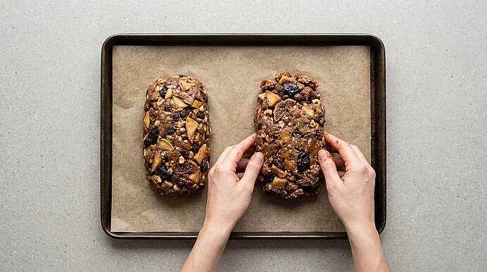 Zwei nussig-fruchtige Müsliriegel auf mit Backpapier belegtem Backblech; Hände fassen das rechte Stück.