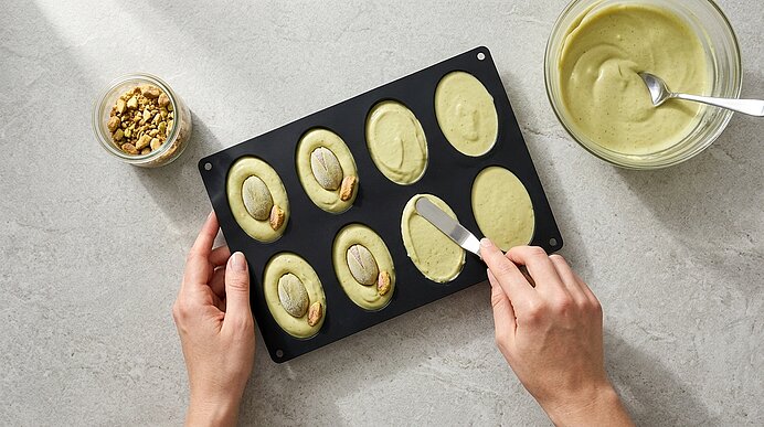 Zwei Hände streichen Pistazienmousse mit einer kleinen Palette in einer schwarzen Silikonform glatt.