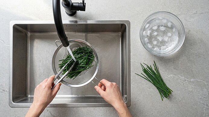 Zwei Hände halten ein Sieb mit Schnittlauch unter das Wasser im Spülbecken. Rechts steht eine Schüssel mit Eiswasser.