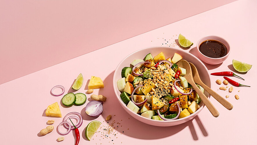 Eine große Schüssel gefüllt mit einem bunten Salat aus Ananas, Gurken, roten Zwiebeln und Erdnüssen. Neben der Schüssel stehen zwei Holzlöffel. Im Vordergrund liegen Limettenstücke und rote Chilis. Dazu gehört eine kleine Schale mit einer dunklen Sauce.