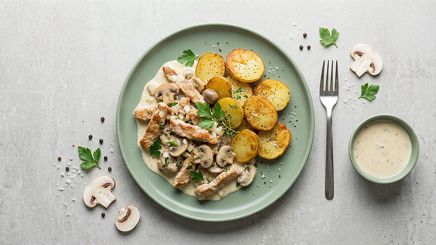 Ein Teller mit zarten Hähnchenstreifen in einer cremigen Sauce, garniert mit frischen Kräutern und Pilzen, daneben knusprige, goldbraun gebratene Kartoffelscheiben. Ein kleines Schälchen mit heller Sauce ist ebenfalls vorhanden.