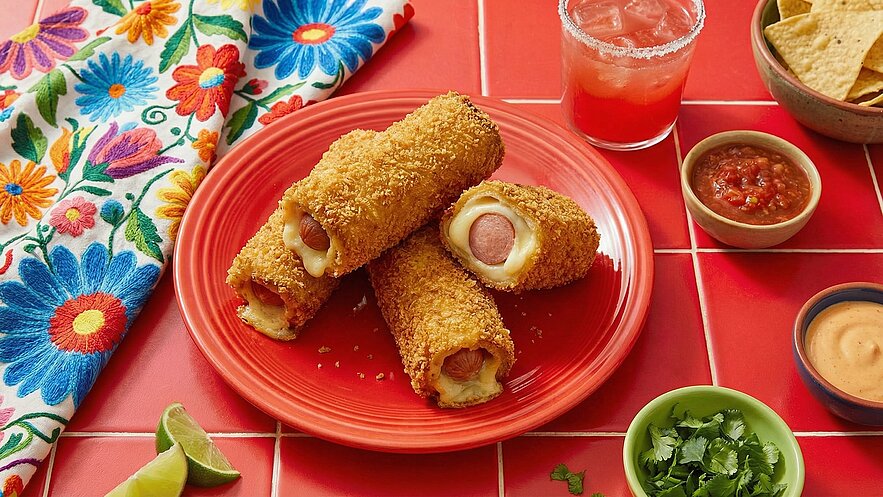 Ein roter Teller mit drei knusprigen Hot-Dog-Rollen, die mit Käse gefüllt sind. Daneben stehen Schalen mit Salsa und Sauce sowie eine Portion Tortilla-Chips. Im Hintergrund befindet sich ein bunt besticktes Tuch.