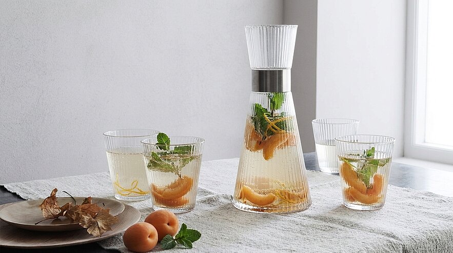Glaskaraffe und drei passende Gläser mit Wasser, Aprikosenscheiben und frischer Minze auf hellem Tischtuch; zwei ganze Aprikosen und kleine Teller daneben.