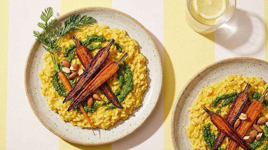 Zwei Teller mit cremigem gelbem Risotto, jeweils garniert mit gerösteten Babykarotten, grünem Kräuterpesto, einigen Nüssen und Karottengrün.