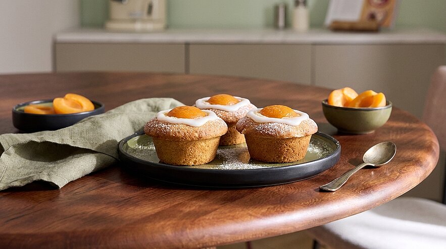 Auf dem runden Holztisch stehen drei Muffins mit Aprikosen und Puderzucker auf einem schwarzen Teller. Neben den Muffins liegt ein grünes Tuch. In zwei Schalen sind geschnittene Aprikosen angerichtet.