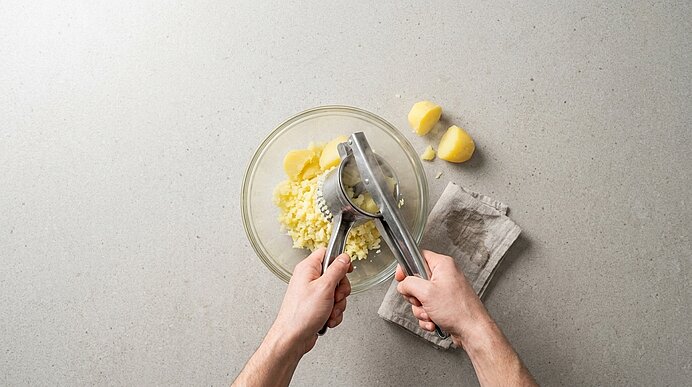 Zwei Hände pressen Knoblauch mit einer Knoblauchpresse über einer Glasschüssel; zerdrückter Knoblauch und zwei geschälte Zehen liegen daneben.
