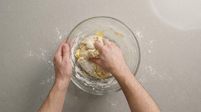 Zwei Hände kneten Teig in einer großen Glasschüssel auf einer grauen Arbeitsfläche, mit etwas verstreutem Mehl.