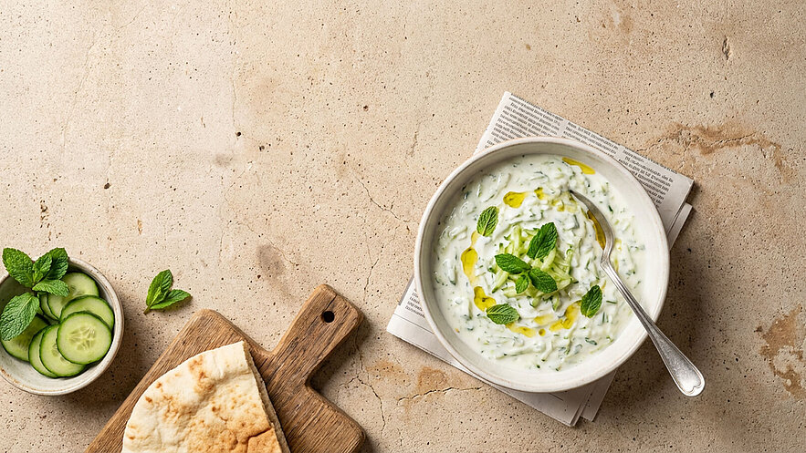 Eine Schüssel mit Gurkenjoghurt, eingelegt in frischen Minzblättern und leichtem Olivenöl. Daneben befinden sich gehobelte Gurkenscheiben in einer kleinen Schale und ein Stück Fladenbrot auf einem Holzbrett. Der Untergrund ist aus hellem Stein.