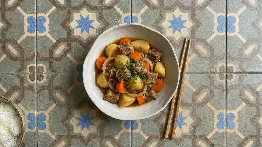 Eine Schüssel mit Fleisch und Gemüse, bestehend aus Kartoffeln, Karotten und Zwiebeln, ist in der Mitte des Bildes. Frische Kräuter garnieren das Gericht. Daneben liegen Essstäbchen aus Holz. Im Hintergrund sind farbige Fliesen sichtbar.