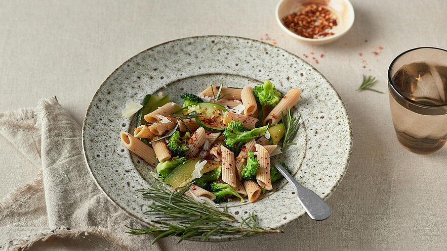 Ein Teller mit Vollkorn-Penne, Garnegeträumt mit Brokkoli und Zucchini. Die Pasta ist mit roten Pfefferflocken und frisch geriebenem Parmesan bestreut, garniert mit Rosmarin. Neben dem Teller steht ein Glas Wasser. Der Hintergrund ist in neutralen Farben gehalten.