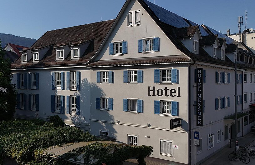 Das Bild zeigt das Hotel Messmer, ein mehrstöckiges Gebäude mit blauen Fensterläden. An der Fassade ist deutlich der Schriftzug "Hotel" zu erkennen. Vor dem Hotel befinden sich einige Bäume und eine kleine Terrasse mit Tischen und Stühlen.